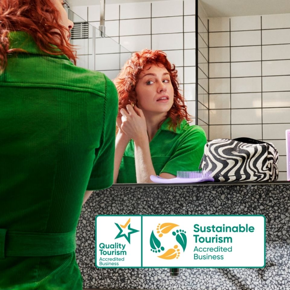 A woman at a bathroom mirror, with Quality Tourism and Sustainable Tourism badges.