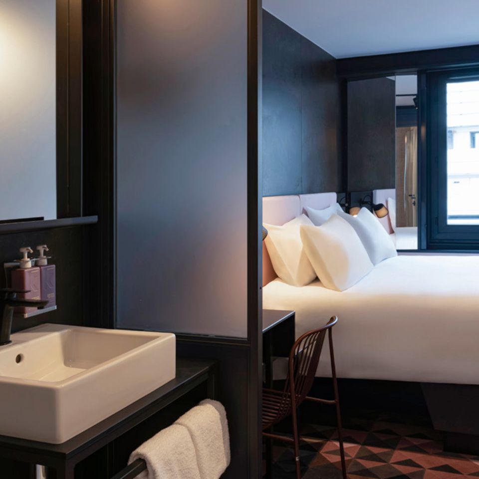 A washbasin with a mirror, in the background is a double bed and a small desk