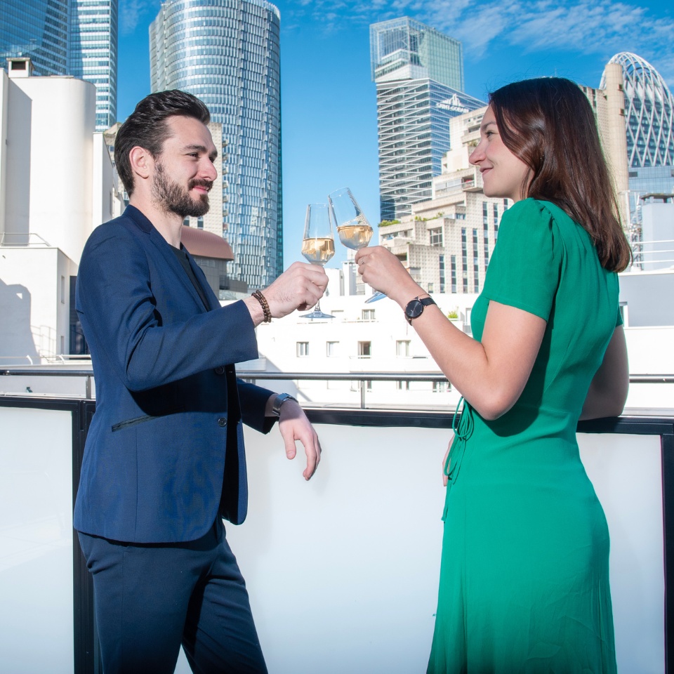 Two people drinking champagne on the balcony, the city sklyine of Paris La Defense is visible.