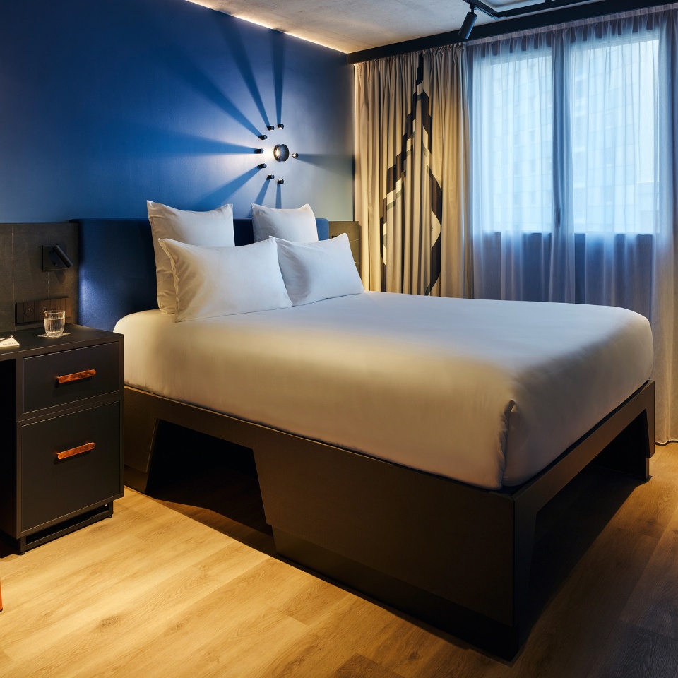 Double bed with white sheets and a blue headboard against a blue wall, to the left is a small black desk