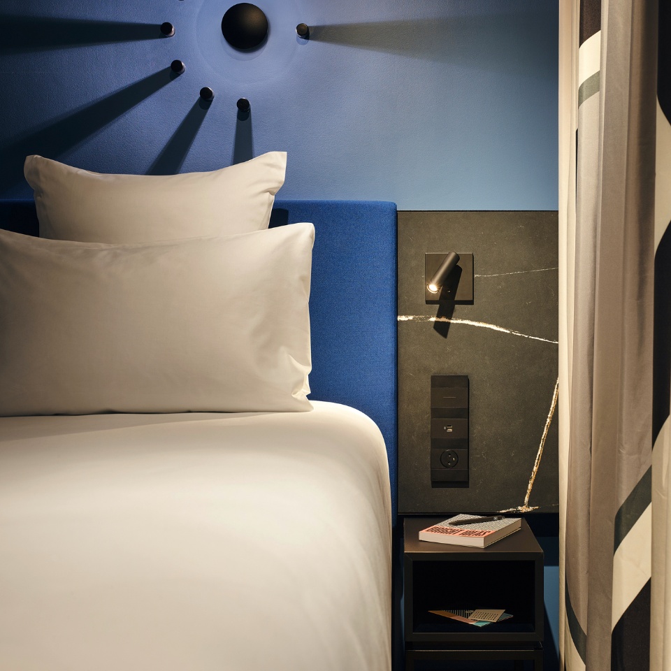 A double bed with a blue headbord, next to it is a black nightstand with a book on it
