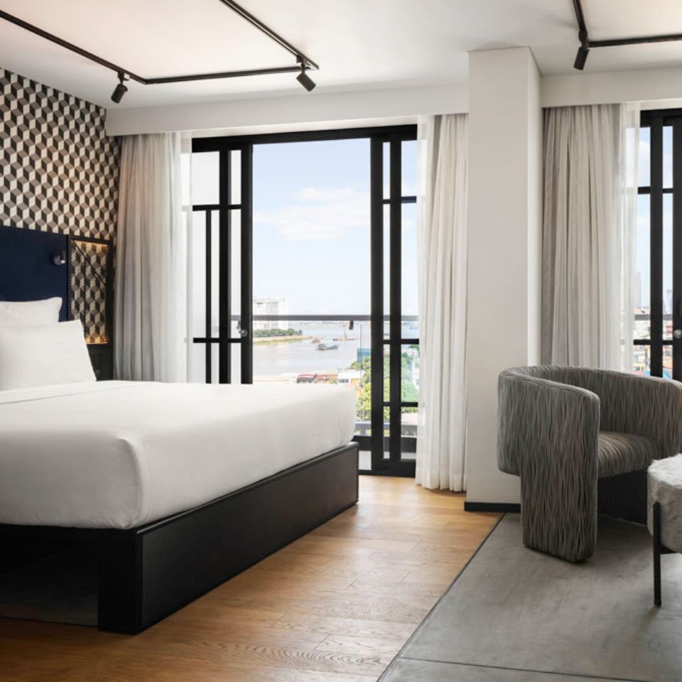 guest room at TRIBE Phnom Penh with a double bed with a blue headboard, a patterned wall, a gray chair and a view of the city