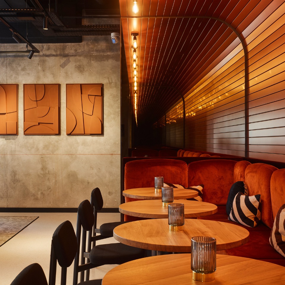 The living space at TRIBE Saint Ouen with several round, wooden tables, black chairs and orange couches