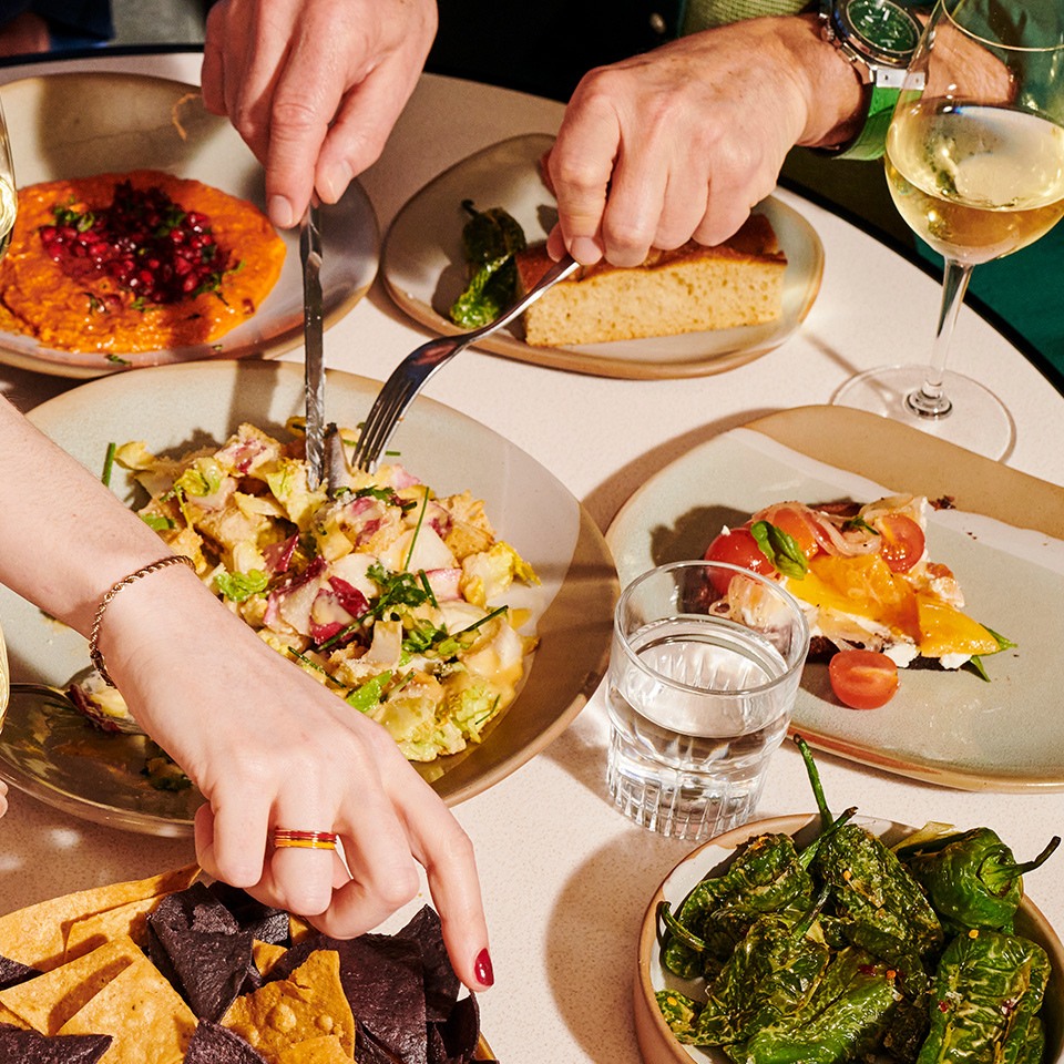 A table set with various dishes, including salad, bread, and appetizers, as two hands serve food while a glass of white wine is nearby.