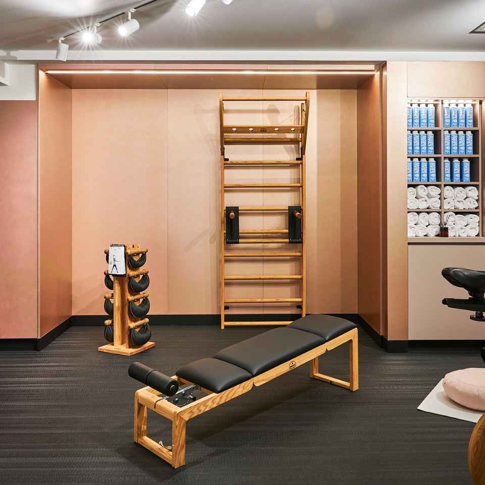 A modern fitness space featuring a wooden exercise bench, a wall ladder, dumbbells, and neatly stacked towels in soft pink tones.