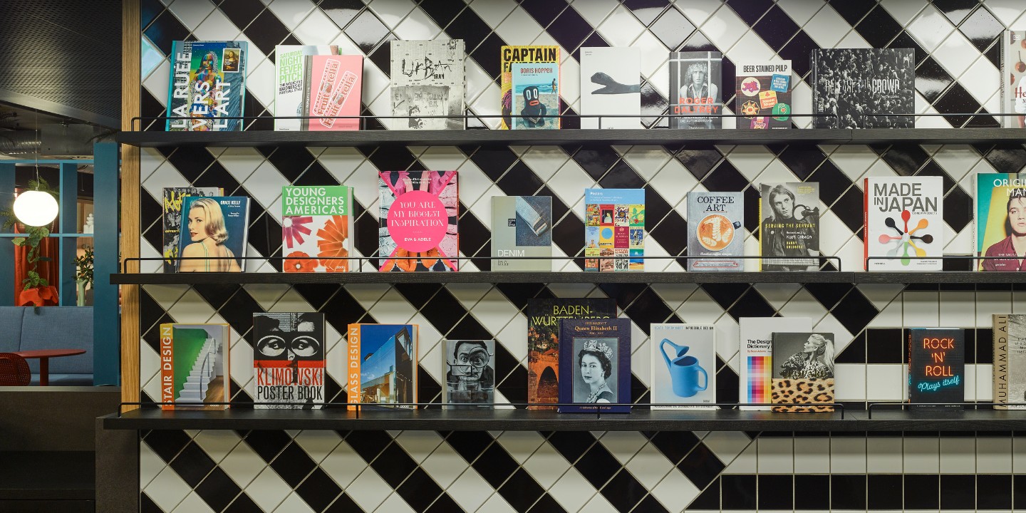A modern bookshelf displaying a variety of colorful books on design, art, and culture, set against a black and white tiled backdrop.