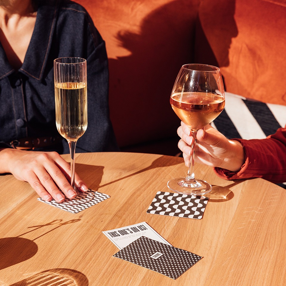 Two people hold drinks above a table with playing cards.