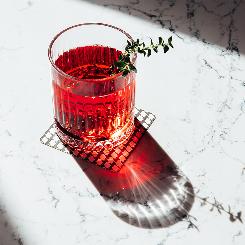 A clear red drink, decorated with some rosemary is sitting on a marble table.