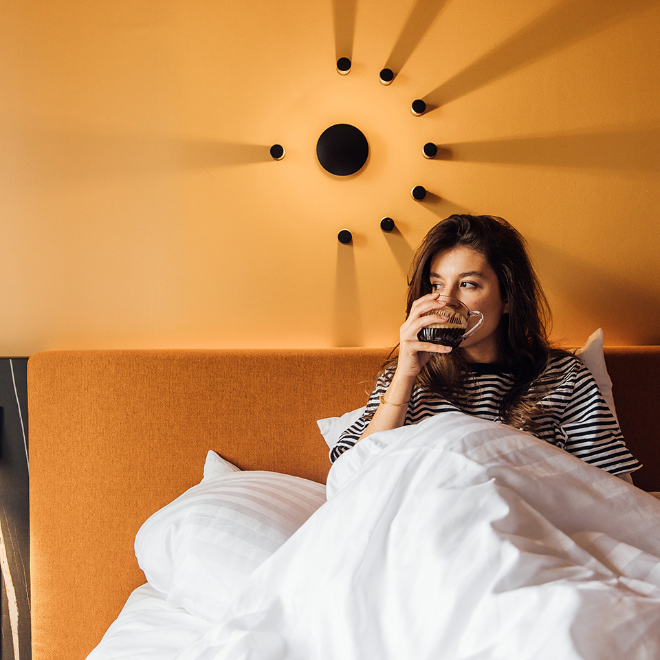 A woman drinks from a cup while sitting in bed against a yellow wall.