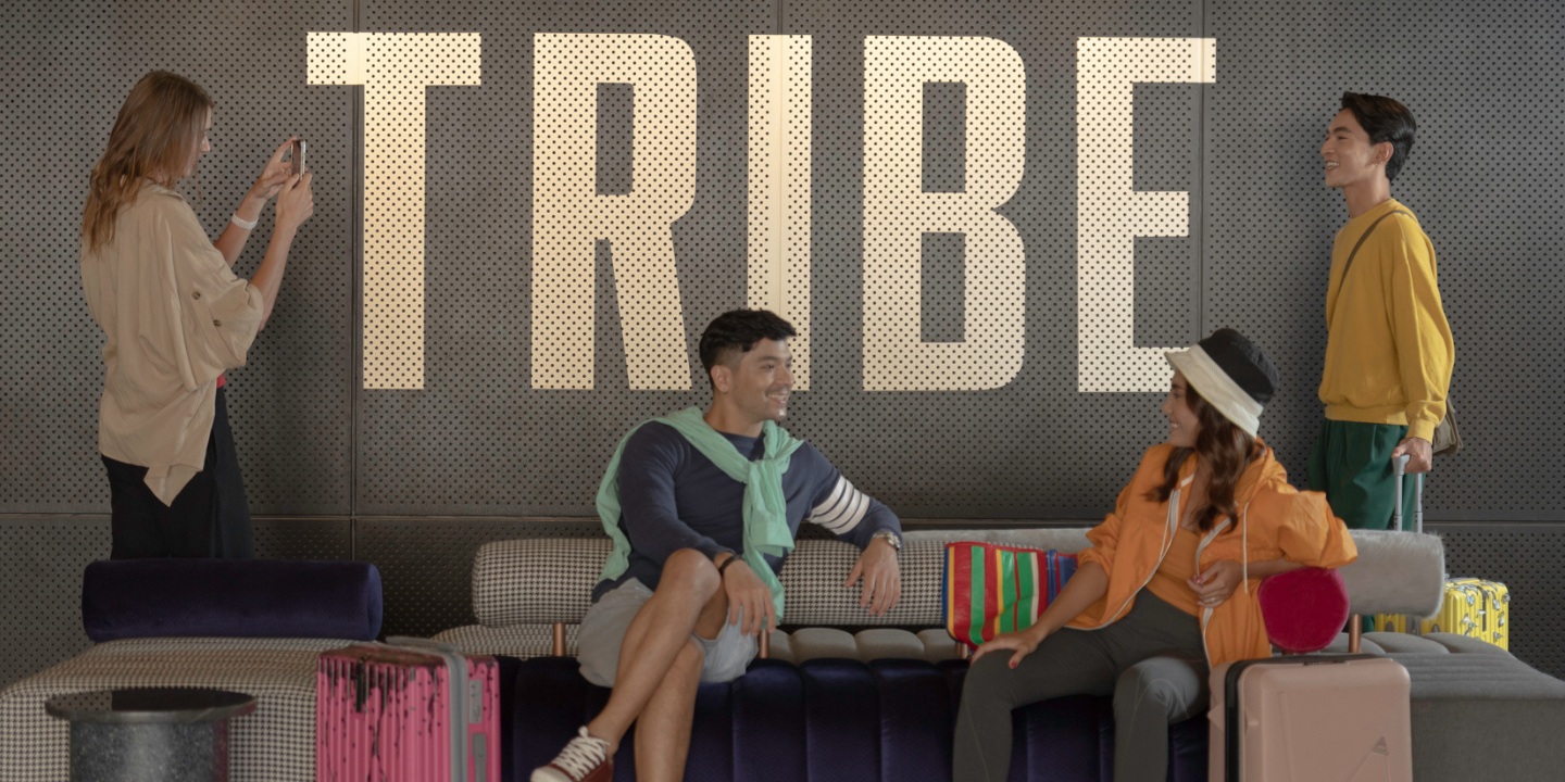 A group of people lounging on sofas with suitcases in head, chatting and smiling. Behind them is a "TRIBE" logo on the wall.
