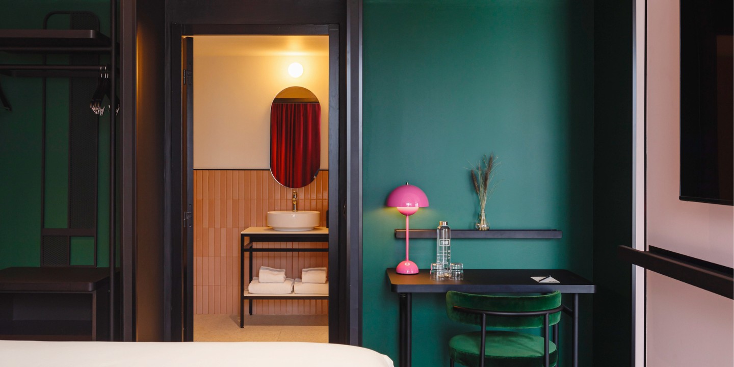 Stylish hotel room featuring a green wall, a pink lamp, and a glimpse of a modern bathroom with a round mirror and towels.