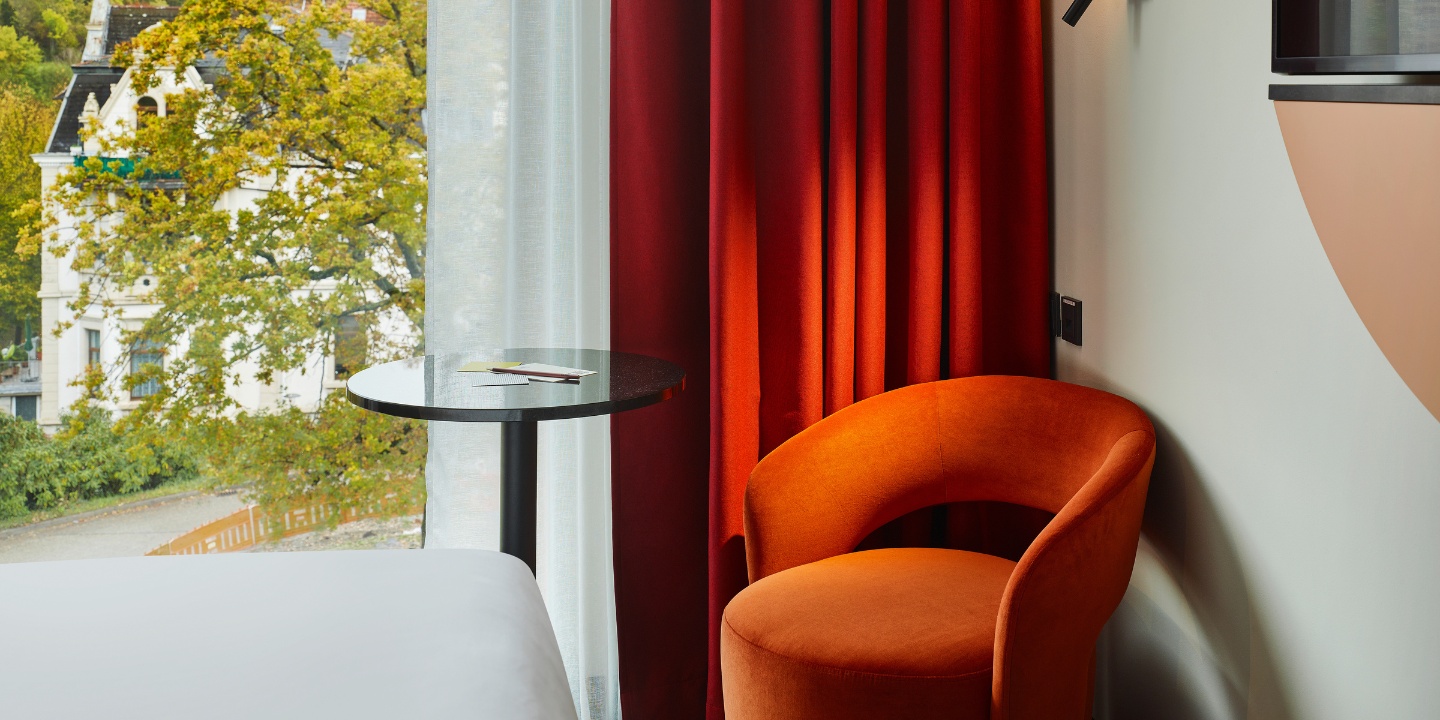 A cozy hotel room features orange accent chair, round table, and red curtains, with a view of greenery outside the window.