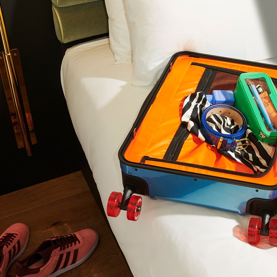 Open suitcase on a hotel bed, packed with clothing and accessories, featuring colorful inner compartments and bright roller wheels.