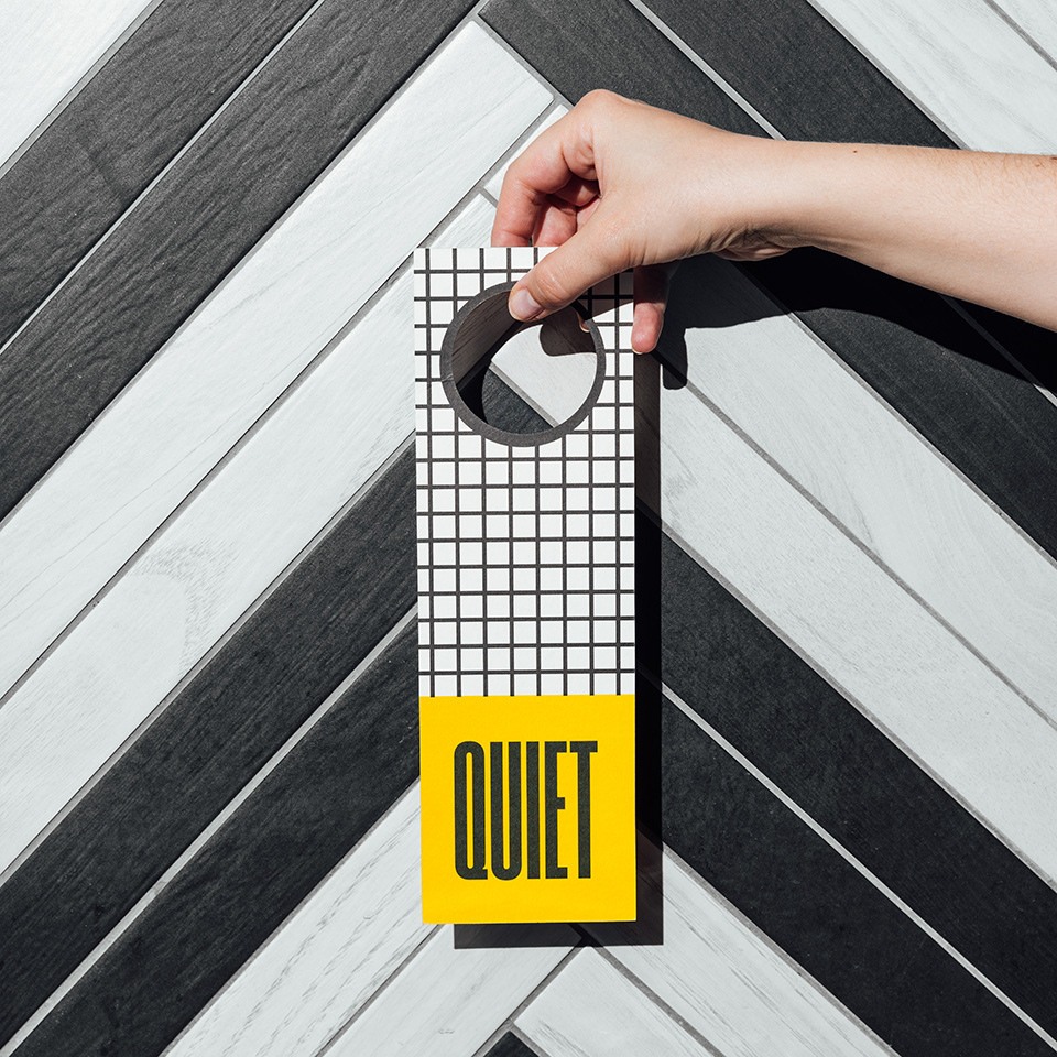 A hand holds a stylish "Quiet" door hanger against a modern black and white herringbone pattern wall.