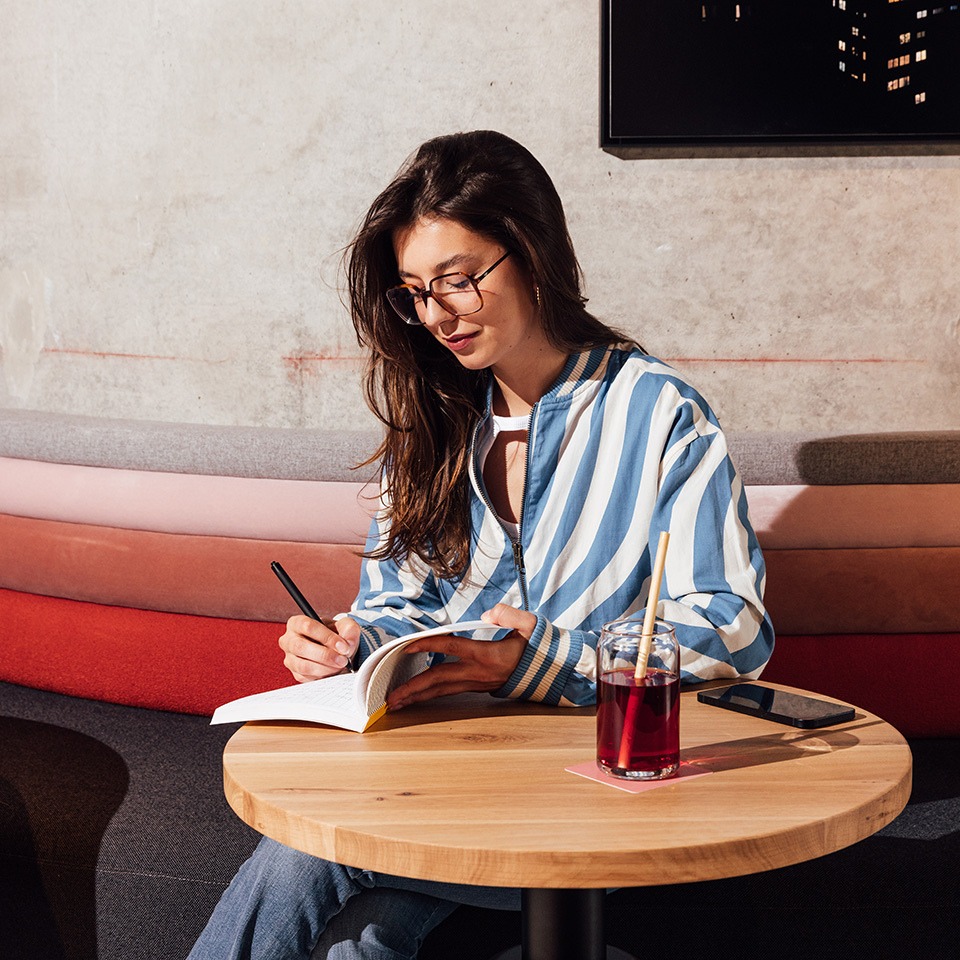 A person in a striped jacket writes in a notebook at a wooden table, with a drink and a smartphone beside them.