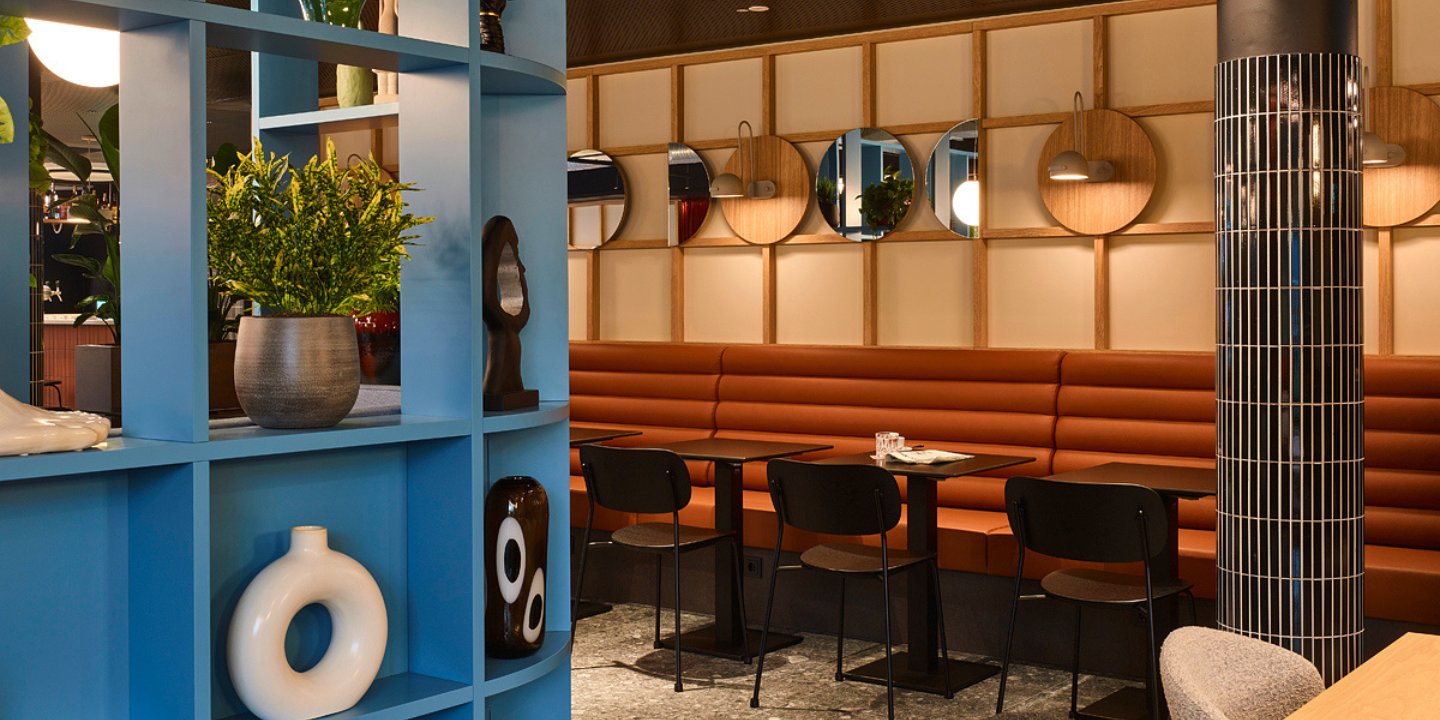 Modern restaurant interior featuring blue shelving, greenery, brown seating, and circular mirrors, creating a stylish dining atmosphere.
