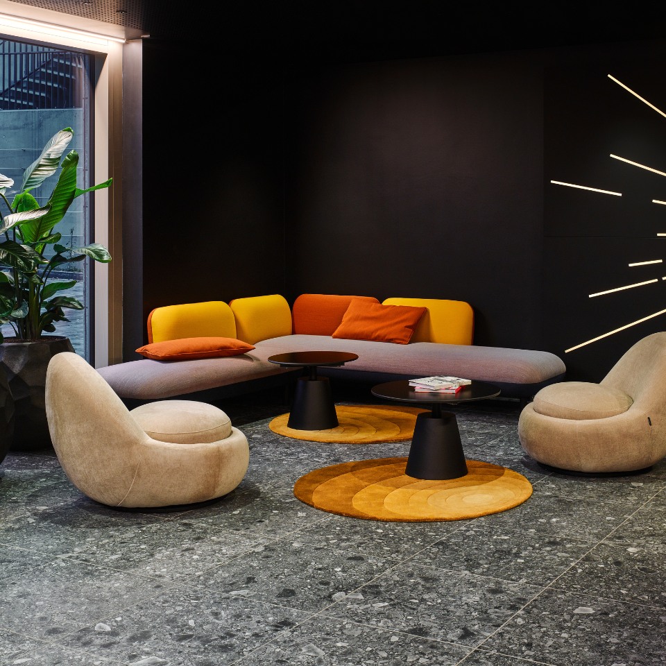 Lounge area with curved sofa, colorful cushions, modern chairs, and circular rugs on a stone floor, enhanced by indoor plants.