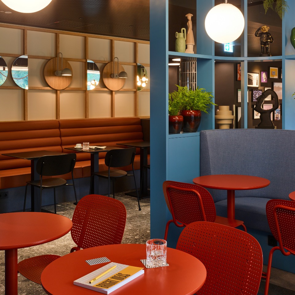Cozy lobby interior with red tables, blue accents, and a mix of seating, featuring decorative plants and contemporary artwork.
