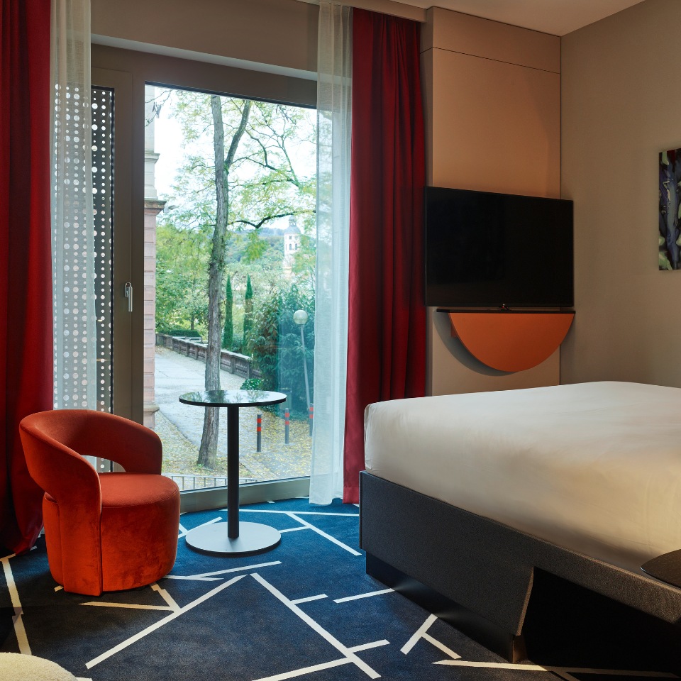 Hotel room featuring an orange chair, round table, plush bed, and large window with red curtains overlooking greenery.