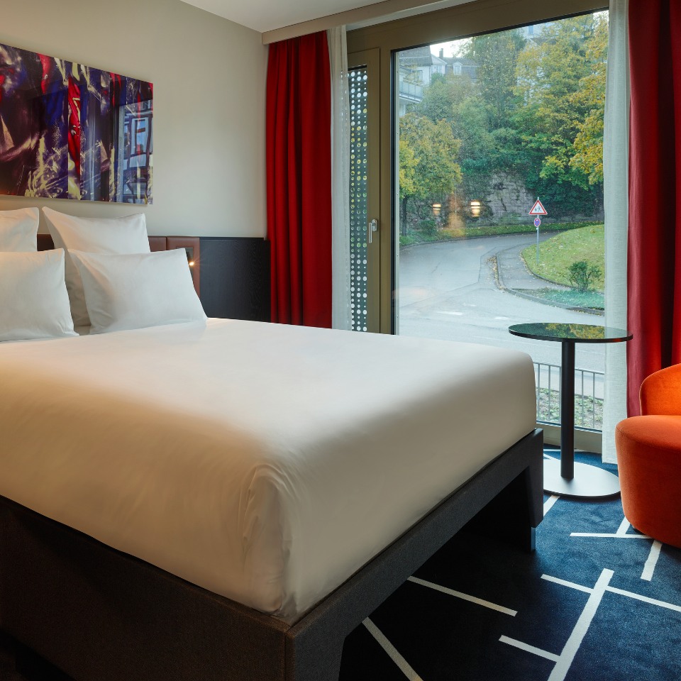 A hotel room featuring a queen bed, vibrant red curtains, an orange armchair, and a large window with a view of the street.