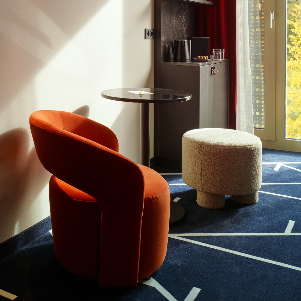 A cozy corner featuring an orange armchair, a round black table, and a textured white stool against a stylish carpet and window backdrop.