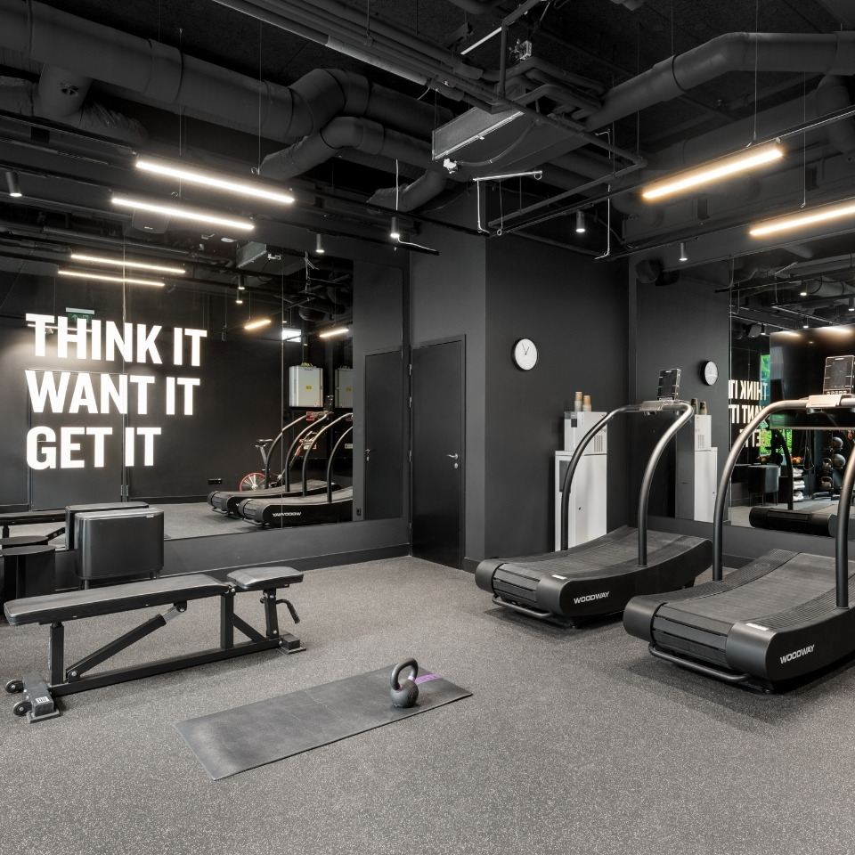 Modern gym interior with treadmills, bench, kettlebell on mat, black walls, and motivational text "Think It Want It Get It" on mirror.