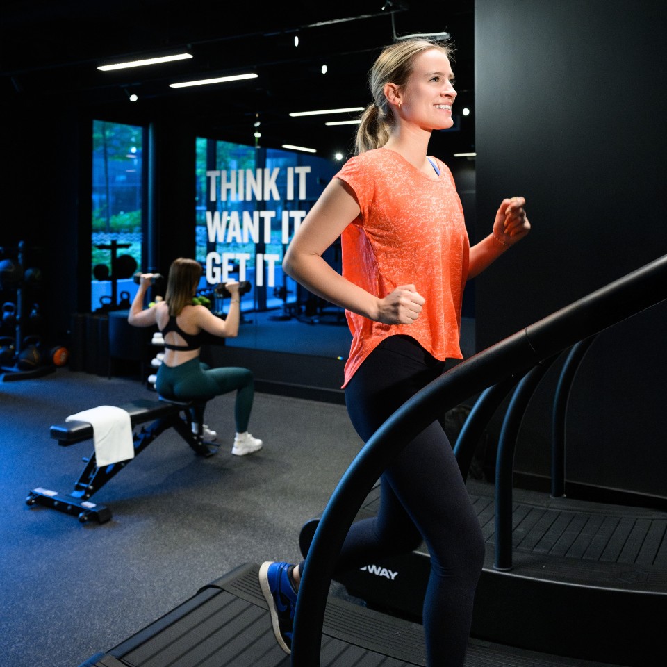 Two women exercising in a gym; one running on a treadmill, the other lifting dumbbells near a bench with motivational words on the wall.