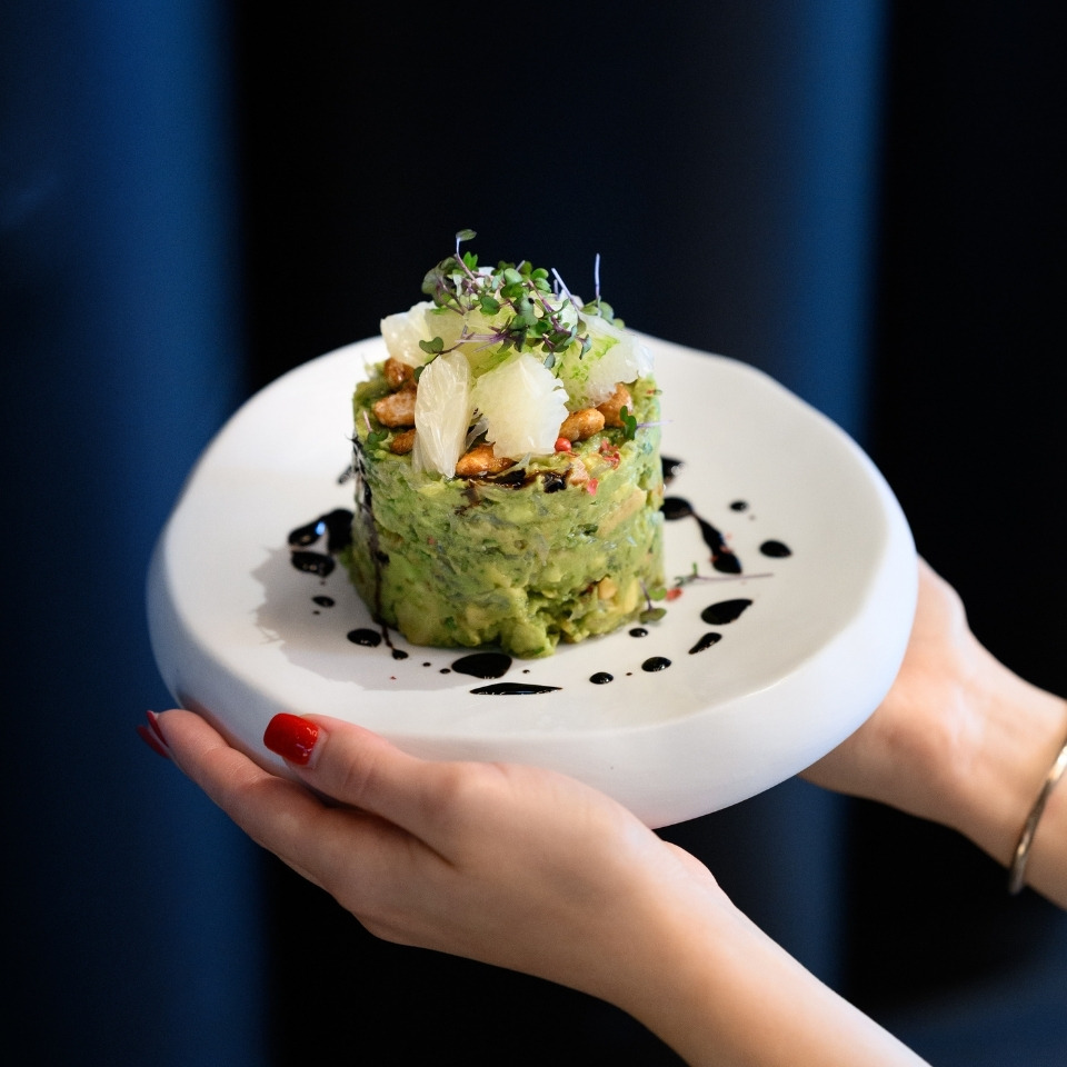 Hands holding a white plate with a gourmet avocado-based dish topped with citrus segments, microgreens, nuts, and a drizzle of dark sauce.