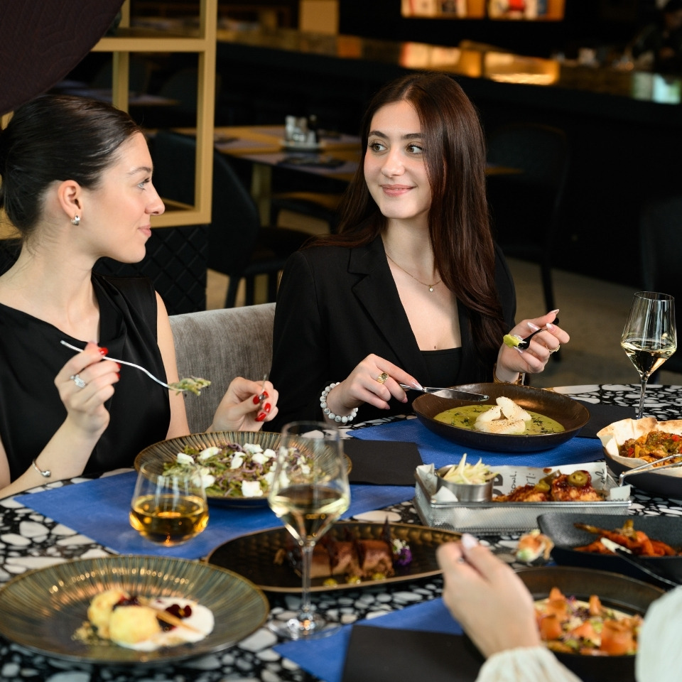 Two women in black outfits enjoying a meal and wine at a restaurant with various plated dishes on the table.