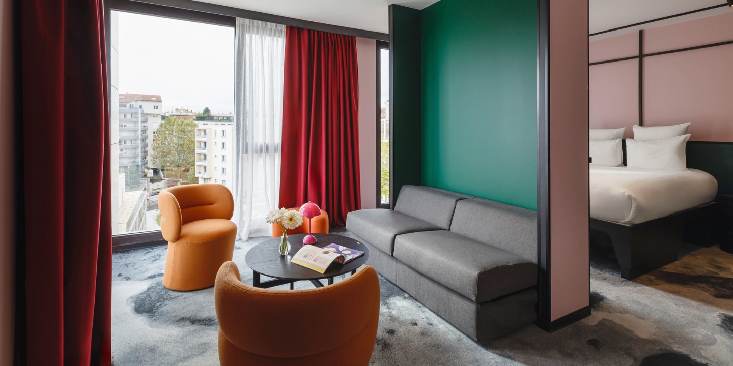 Modern hotel room featuring a gray sofa, orange chairs, colorful walls, and large windows with city views. Cozy and stylish atmosphere.