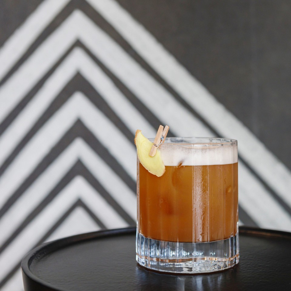A glass of cocktail with amber liquid, topped with foam and a lemon slice, resting on a black table against a muted backdrop.