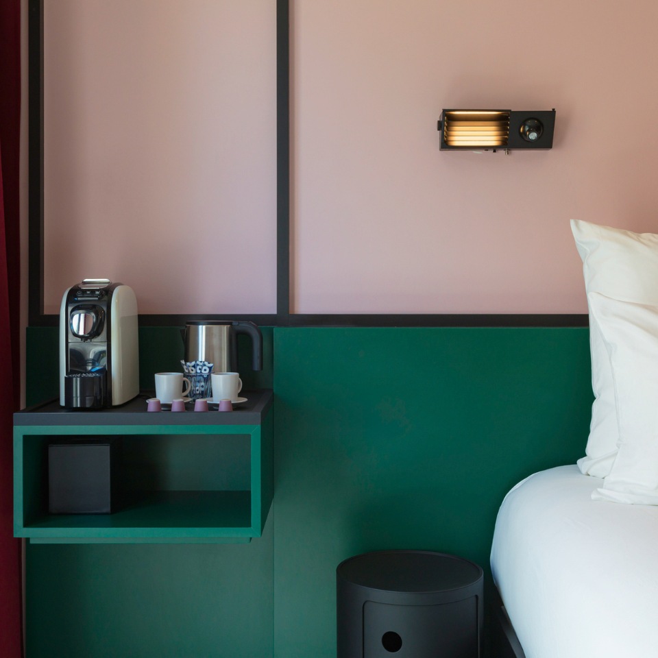 Modern hotel room corner featuring a green coffee station, cup set, and stylish wall light against a pink background.
