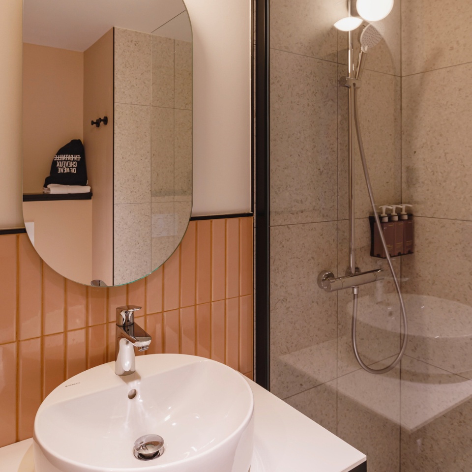 Modern bathroom featuring an oval mirror, a sleek sink, warm orange tiles, and a stylish shower with toiletries on display.