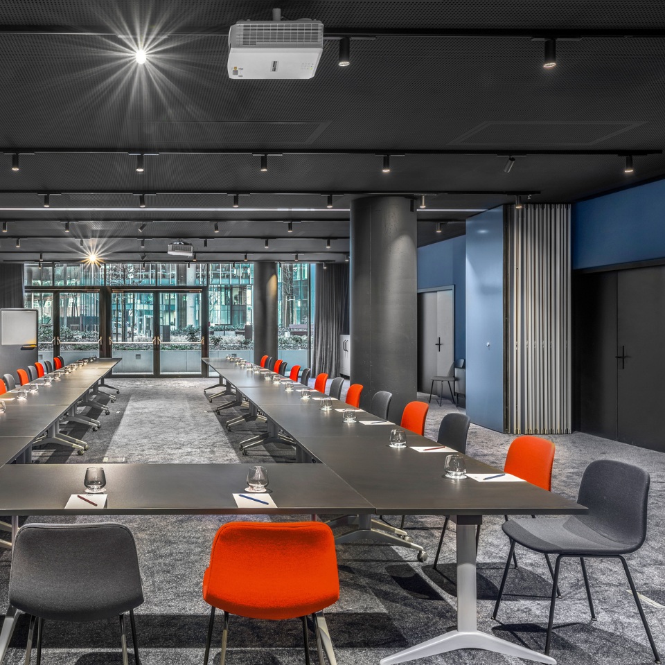 Conference room with long tables, colorful chairs, and a projector, illuminated by overhead lights and natural light through large windows.
