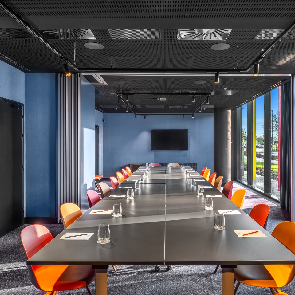 Conference room with a long table, orange chairs, and a large screen on a blue wall, brightened by natural light.