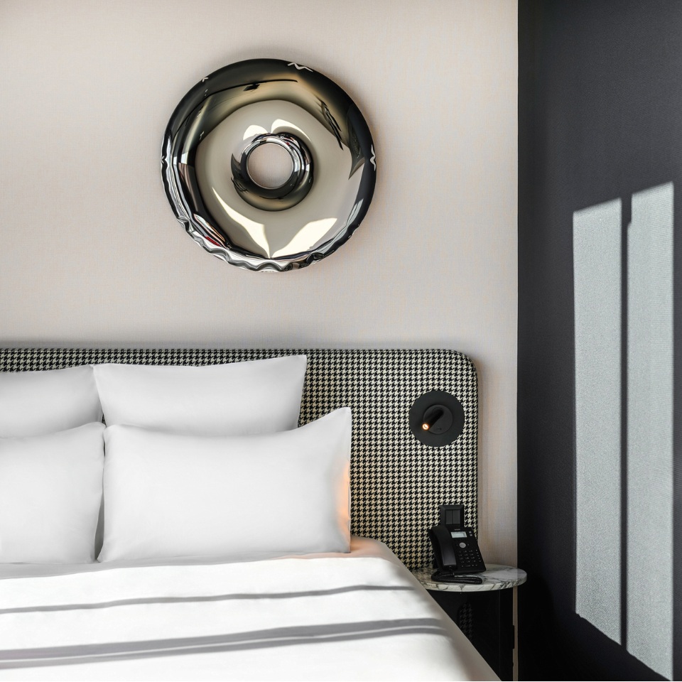 A modern hotel room featuring a stylish bed with white pillows, a houndstooth headboard, and a circular wall decor piece above.