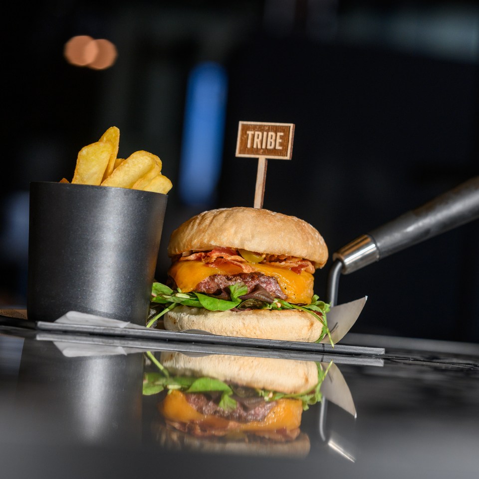 A juicy cheeseburger with bacon, lettuce, and pickles, accompanied by a cup of crispy fries, served on a reflective surface.