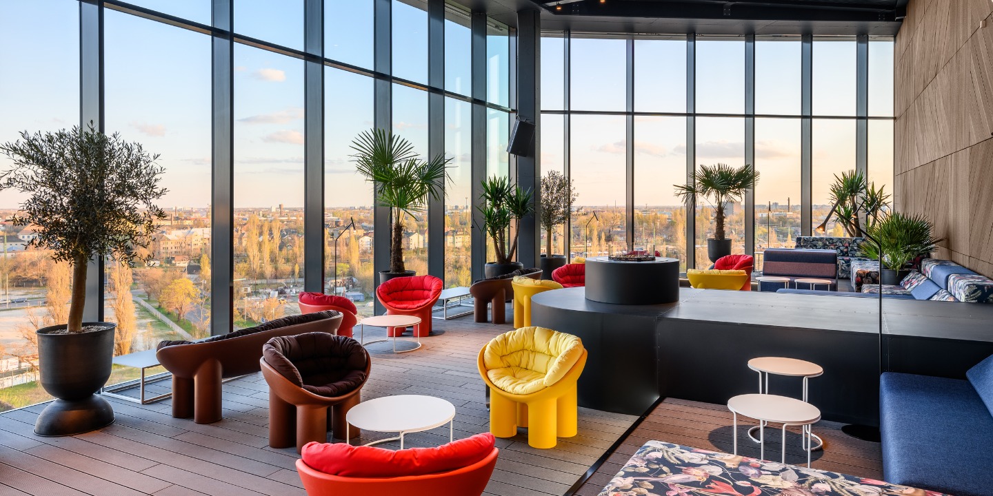 A stylish var with colorful seating, large windows, and a panoramic city view, adorned with plants and modern decor.