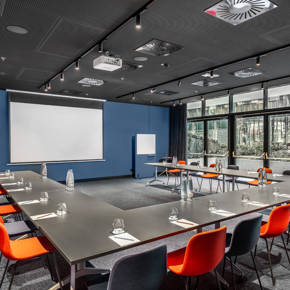Salle de conférence moderne avec une table en U, des chaises orange, un écran de projection et de grandes fenêtres donnant sur la verdure.