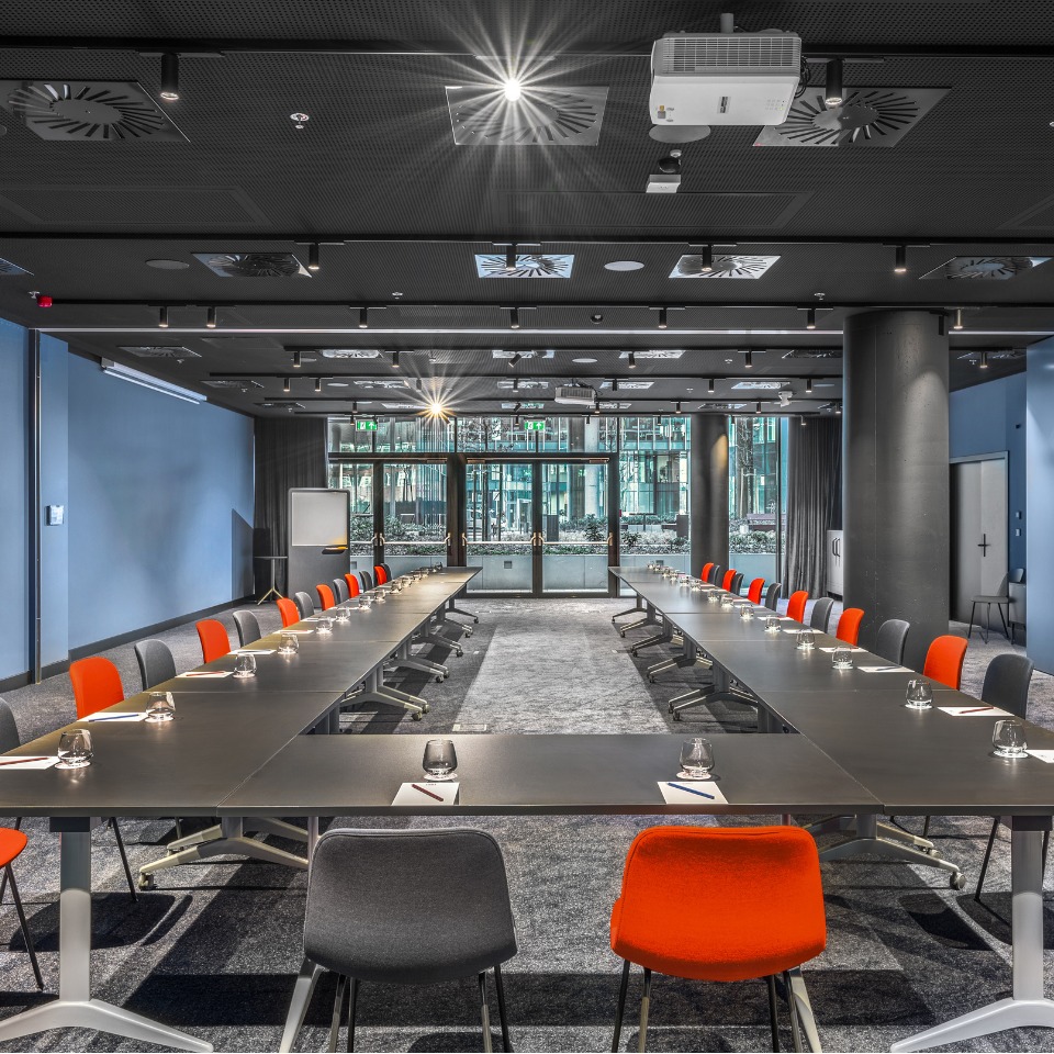 Salle de conférence moderne avec de longues tables disposées en U, des chaises colorées et de grandes fenêtres laissant entrer la lumière naturelle.