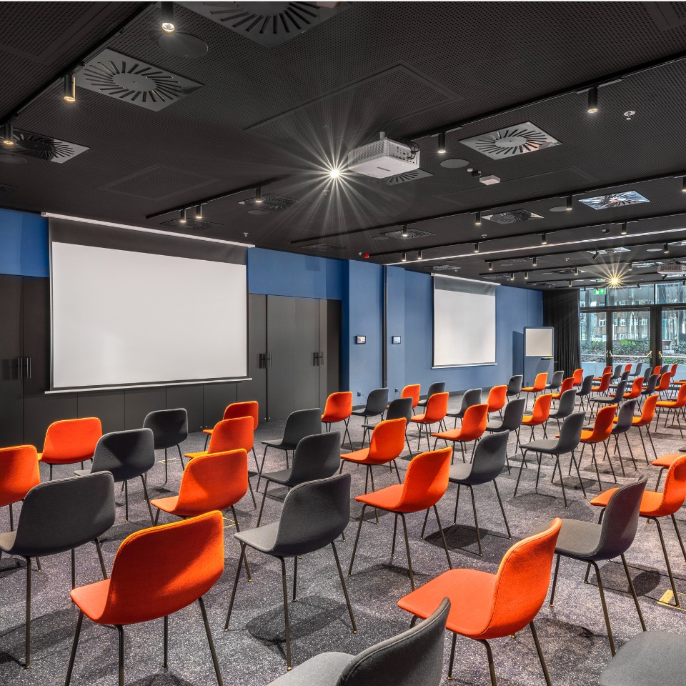 Une grande salle de conférence avec des rangées de chaises noires et orange, deux écrans de projection et de grandes fenêtres laissant entrer la lumière naturelle.