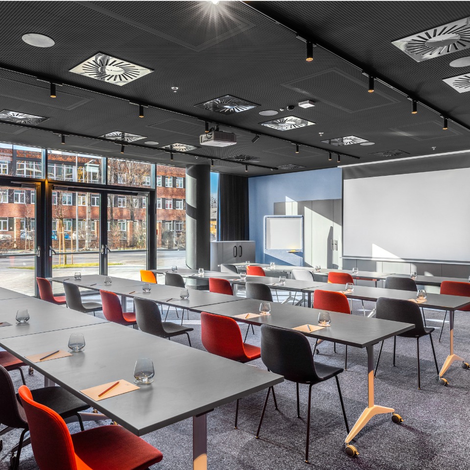 Salle de conférence avec des tables grises, des chaises colorées et un grand écran, baignée de lumière naturelle par les fenêtres.