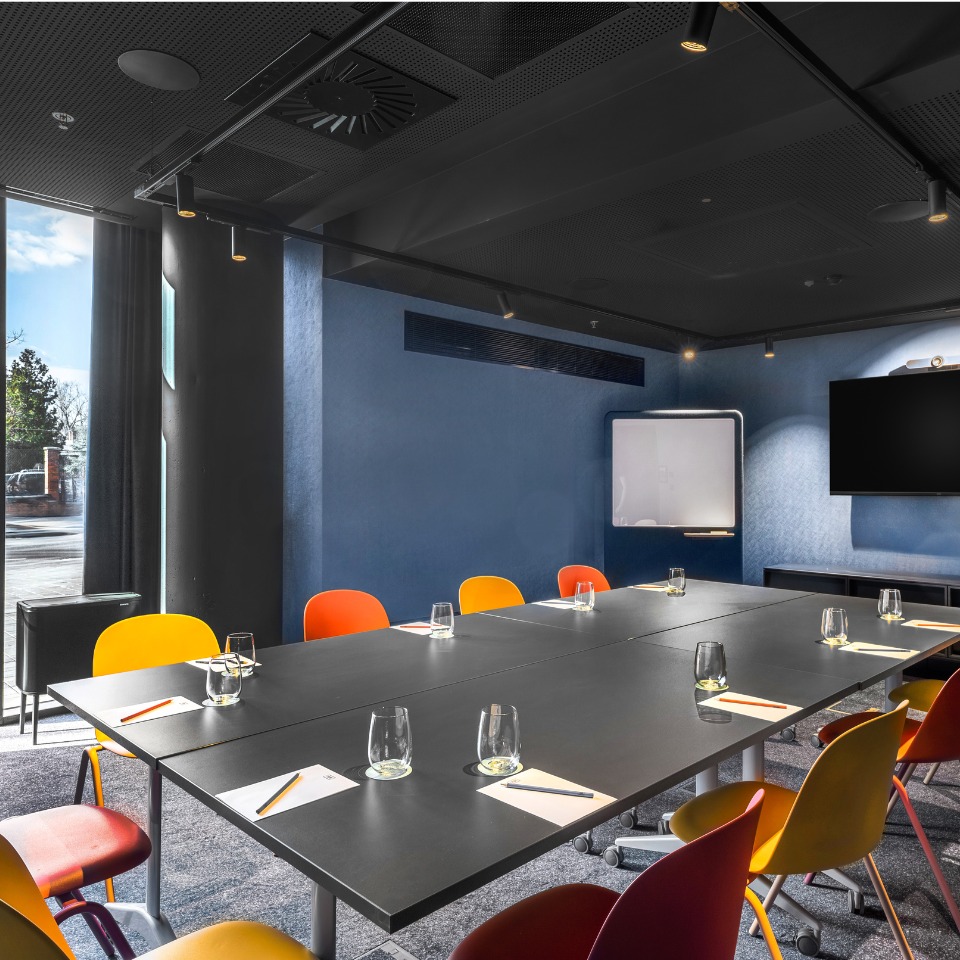 Une salle de conférence avec une longue table, des chaises colorées, des verres d’eau, un tableau blanc et un écran de télévision.