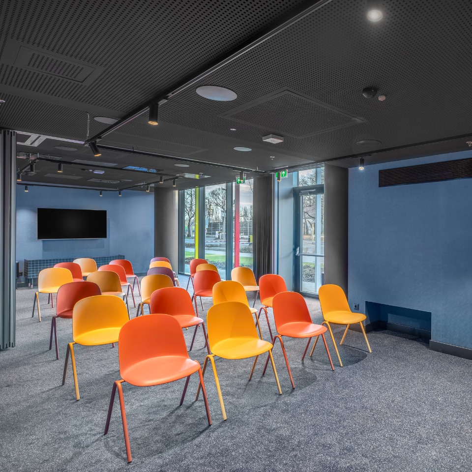 Une salle de conférence avec des chaises orange vif et jaunes disposées en rangées, de grandes fenêtres et une télévision murale.