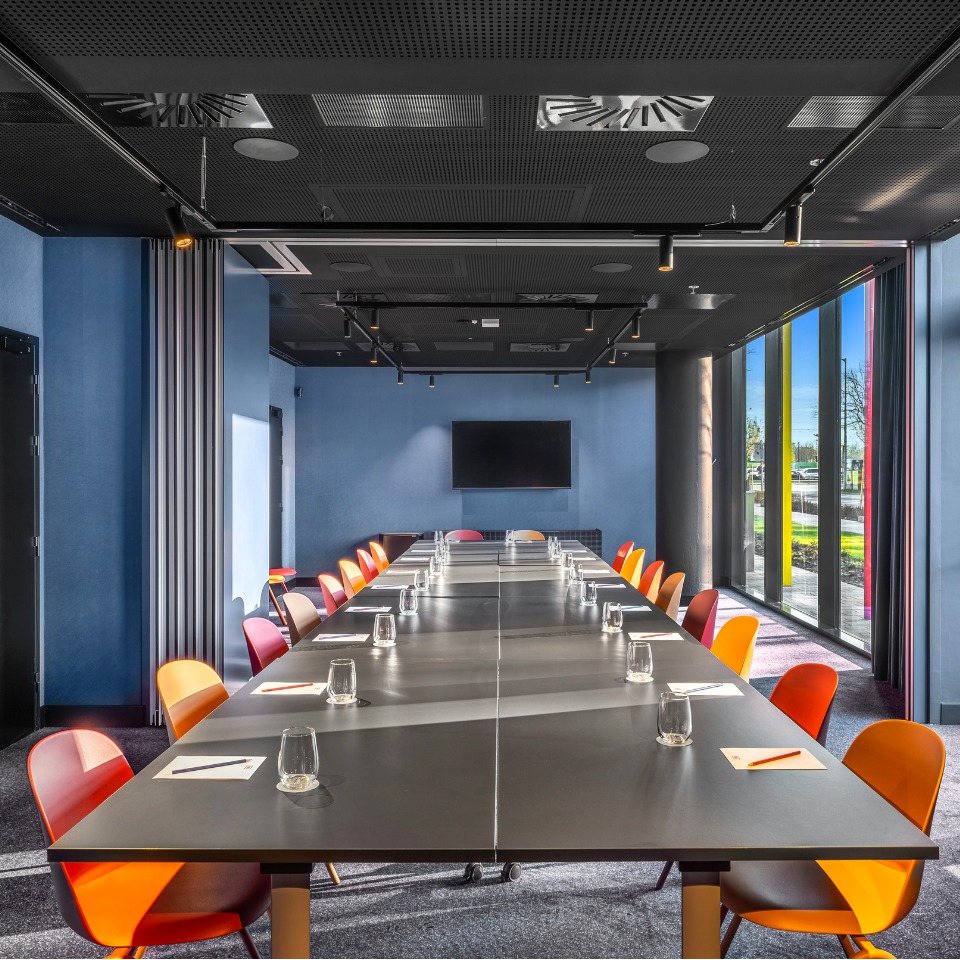 Salle de conférence avec une longue table, des chaises colorées et un grand écran, baignée de lumière naturelle grâce à des baies vitrées du sol au plafond.