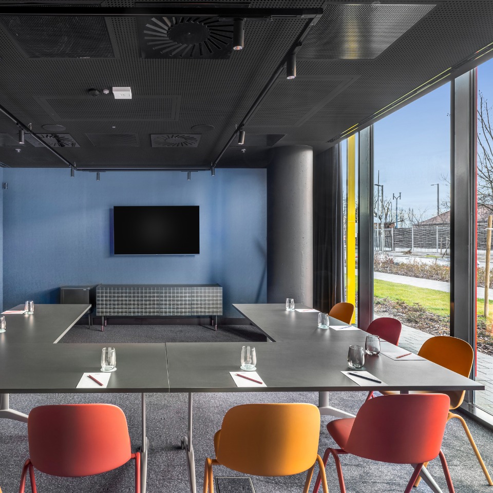 Salle de conférence au plafond sombre, aux murs bleus, avec un écran de télévision, des chaises colorées autour d’une table épurée et de grandes fenêtres.