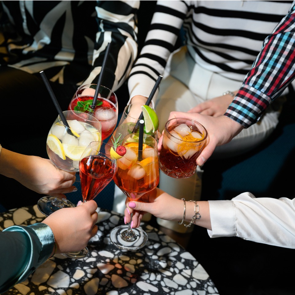 A group of hands raising colorful cocktails in celebration, featuring various drinks with fruits and ice, on a stylish table.