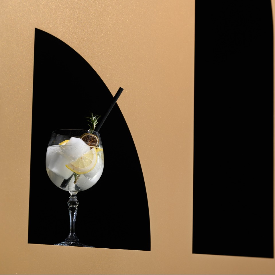 A refreshing cocktail in a glass with ice, lemon slices, and a black straw, set against a gold and black backdrop.