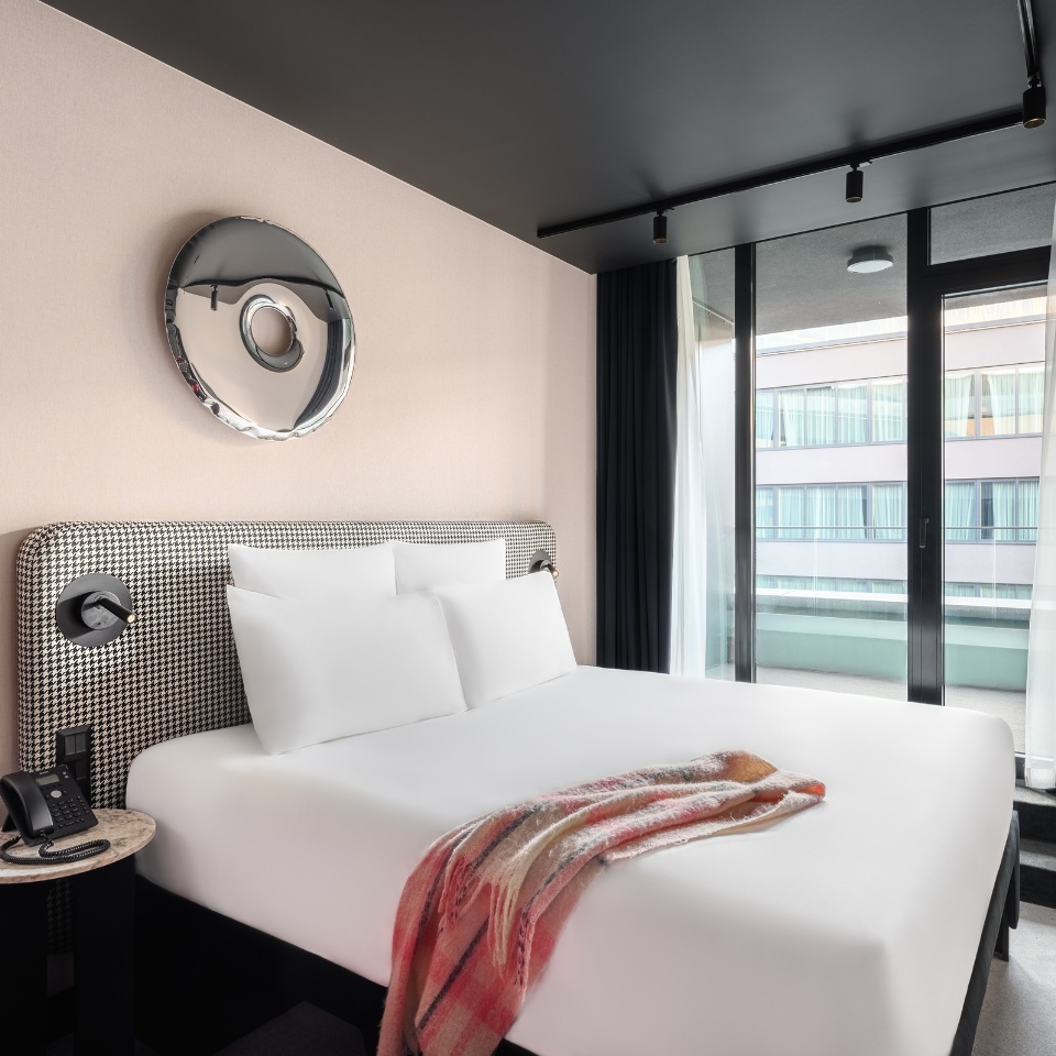 Chambre d’hôtel avec un lit élégant, un miroir décoratif et des portes-fenêtres donnant sur un balcon, habillées de voilages, rehaussée d’un plaid douillet.