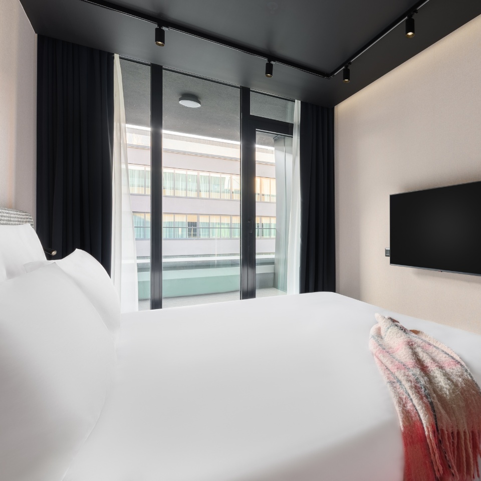 Chambre d’hôtel avec un lit King size, une décoration épurée et de grandes fenêtres donnant sur un balcon. Télévision murale.